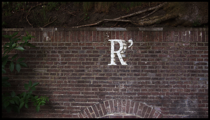 A capital R with a right single quote painted on a brick wall. class=